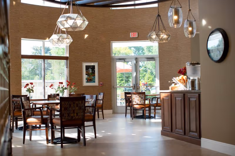 A bright dining area with several wooden tables and chairs, each table decorated with a vase of red flowers. Large windows and glass doors allow natural light to fill the space, showing a view of greenery and outdoor umbrellas. Modern pendant lights hang from the ceiling, and a wooden sideboard with a water dispenser and cups is visible on the right side of the image.