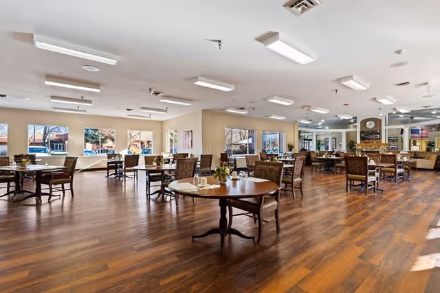 Spacious dining room with multiple round and square tables, each set with chairs and small flower arrangements. Large windows allow natural light to fill the room, and the floor is covered with wood-style flooring. The room has a bright and welcoming atmosphere.