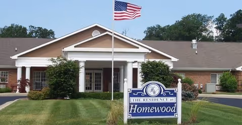 Front exterior view of a single-story brick building with white columns at the entrance, an American flag on a flagpole in front, and a sign that reads 'The Residence at Homewood' on a well-maintained lawn.