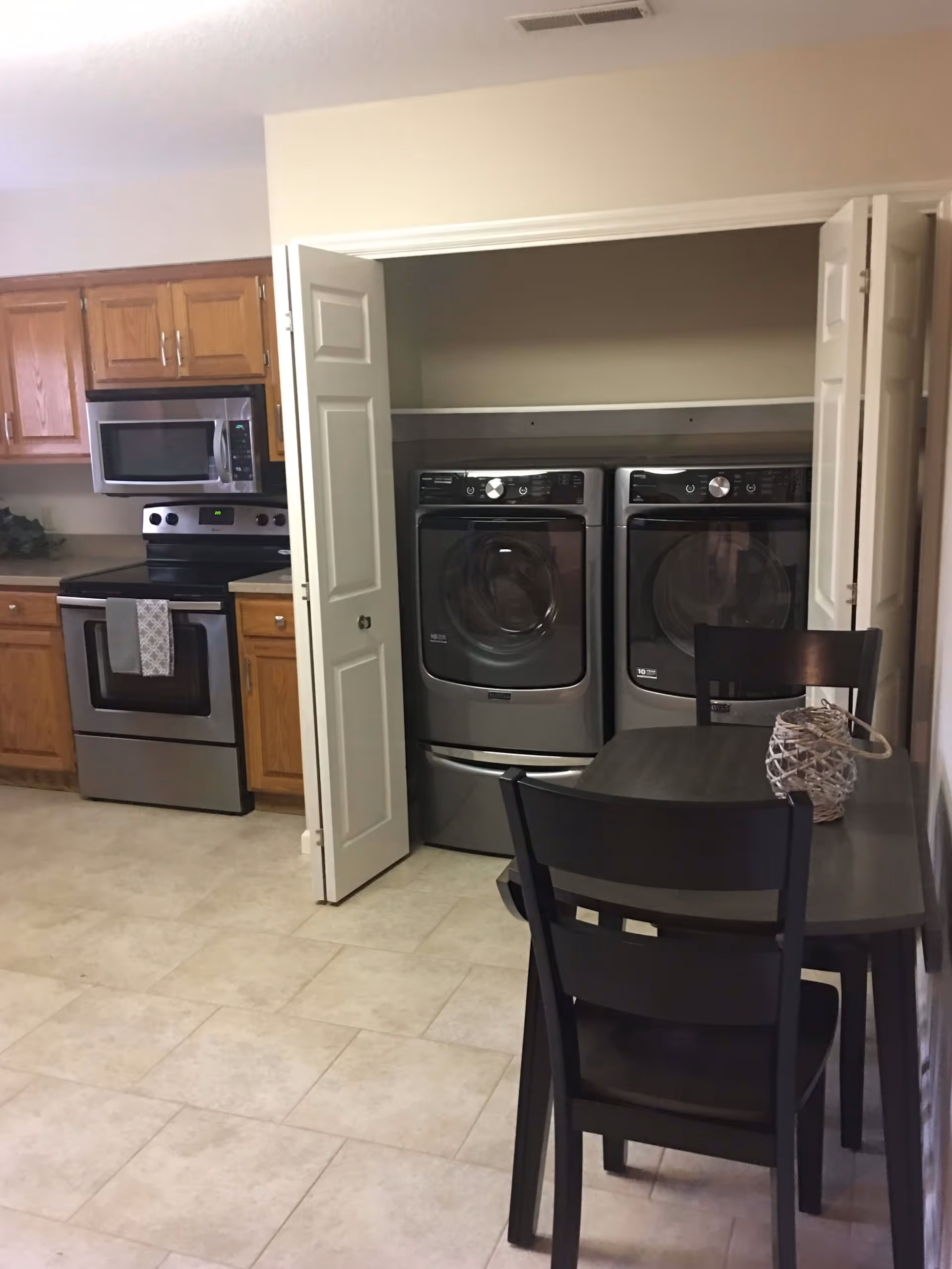 Interior view of a kitchen area with wooden cabinets, a stainless steel microwave and stove, and a small dining table with two black chairs. To the right, there is a laundry closet with white bi-fold doors open, revealing a front-loading washer and dryer.