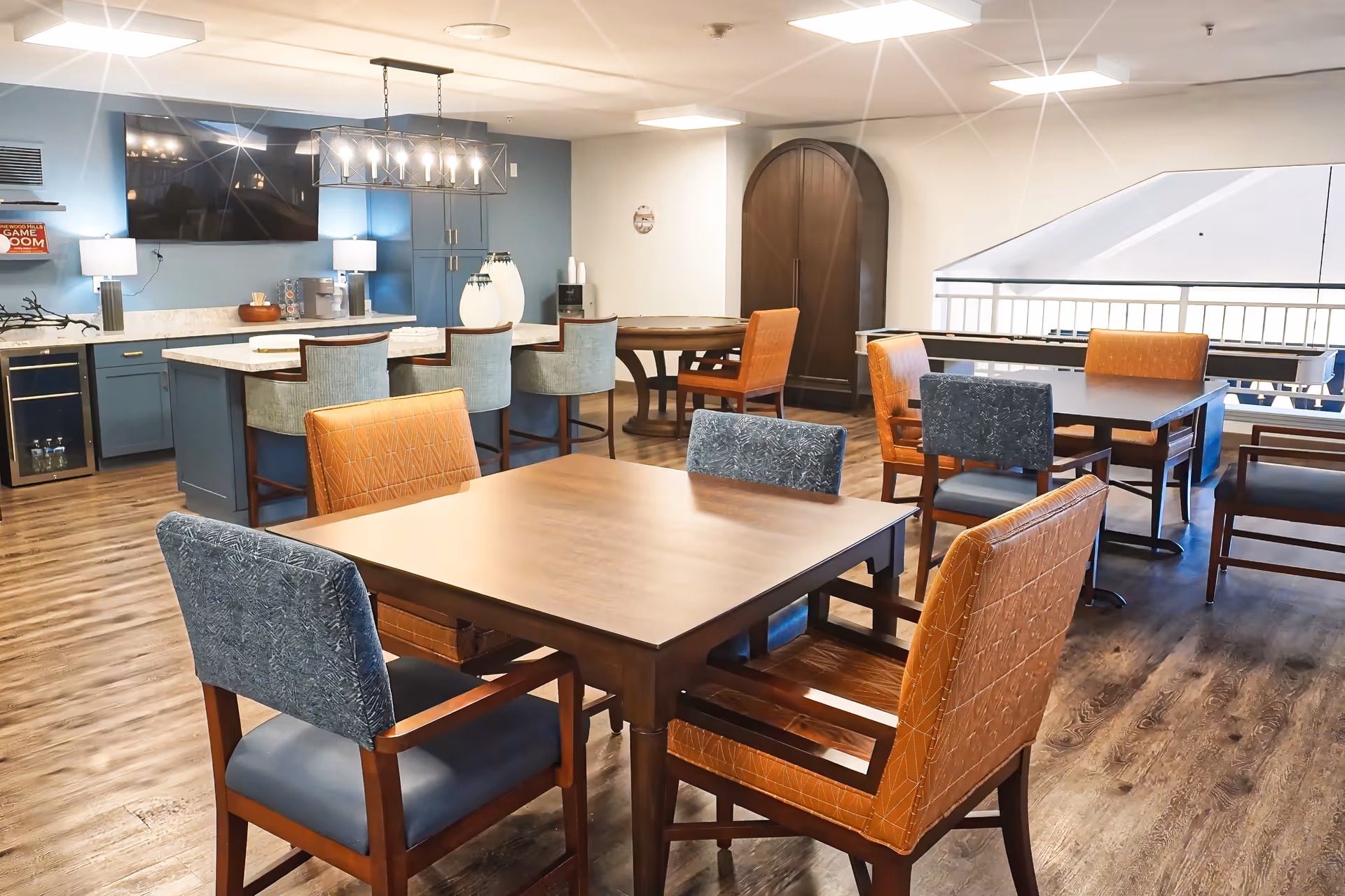A bright and modern dining area in a senior living facility featuring wooden tables with mixed upholstered chairs in blue and orange. In the background, there is a kitchen island with bar stools, a large flat-screen TV mounted on a blue accent wall, and cabinetry. The flooring is wood-style, and the space is well-lit with ceiling lights and lamps.