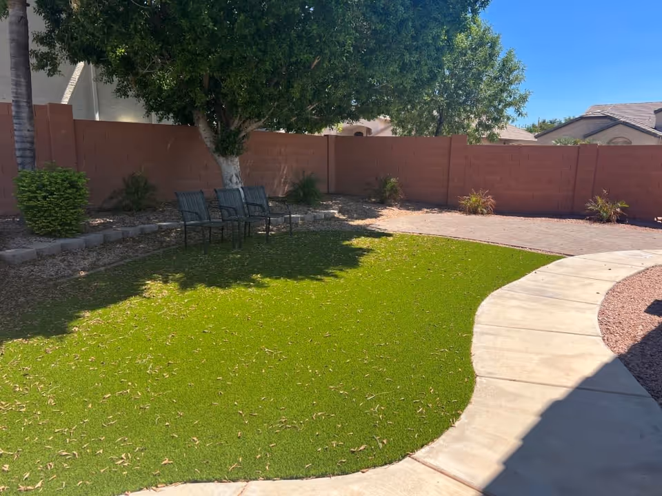 Outdoor area with a curved concrete pathway, green grass lawn, three metal chairs under a tree, and a brown brick wall enclosing the space. Some small plants and shrubs are along the wall, with a clear blue sky overhead.