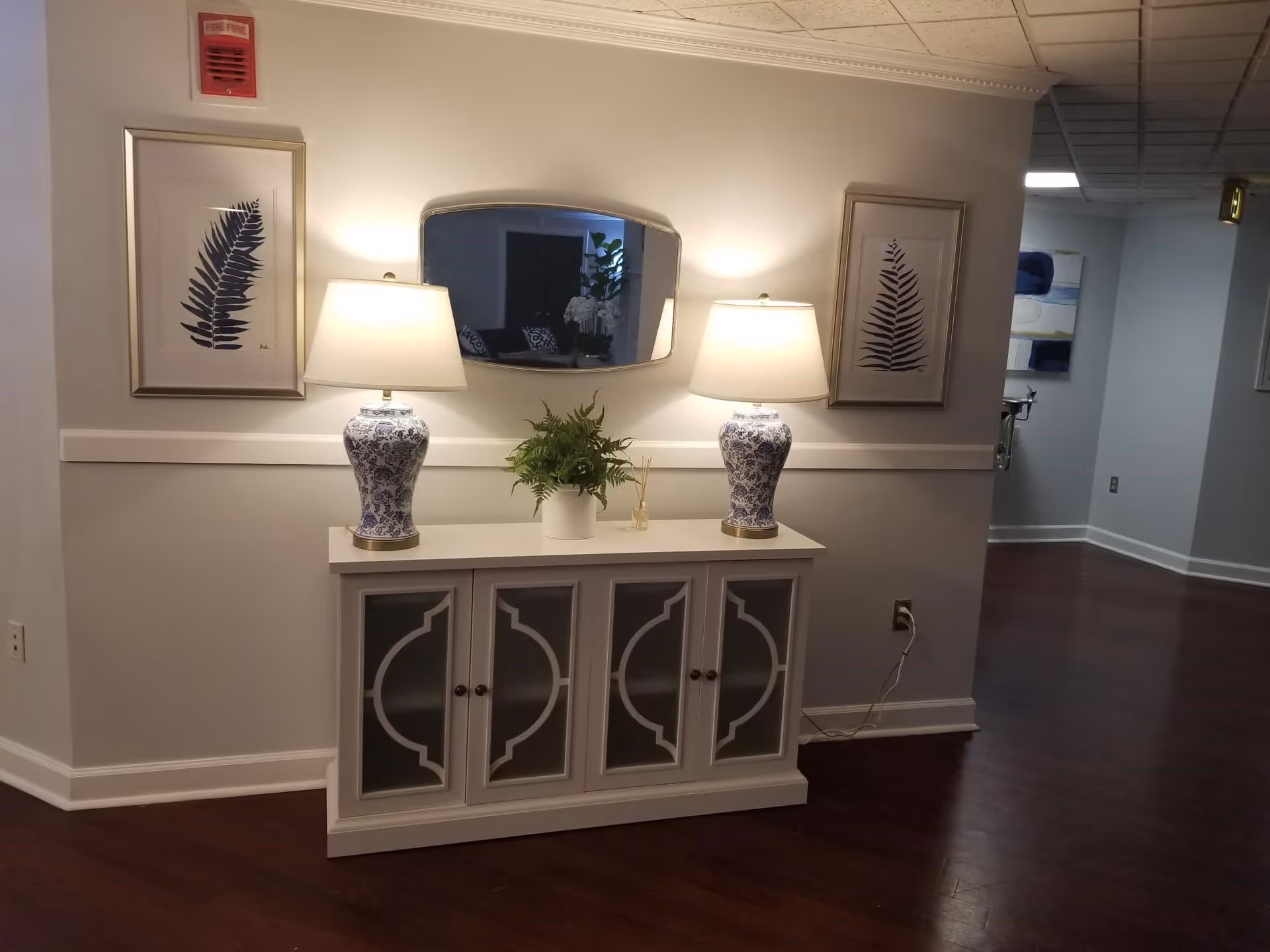 A hallway corner in a senior living facility with a white cabinet topped by two blue and white patterned lamps and a small green plant in the center. Above the cabinet is an oval mirror flanked by two framed botanical prints. The floor is dark wood, and the walls are painted light gray.
