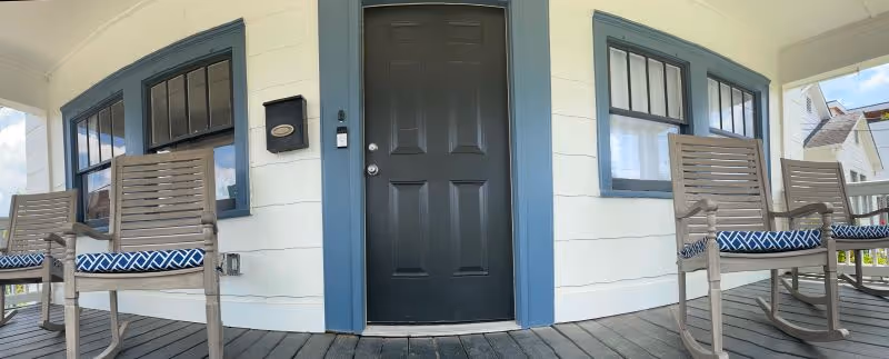 Front porch of a house with a black door framed by blue trim, two windows with blue trim on either side, and four wooden rocking chairs with blue and white patterned cushions placed on a wooden floor.