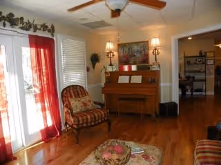 A cozy living room with hardwood floors featuring a piano against the wall, a cushioned armchair with a striped pattern, a ceiling fan with lights, and red curtains covering glass doors. There is a small table with a decorative centerpiece in the foreground and a hallway leading to another room in the background.