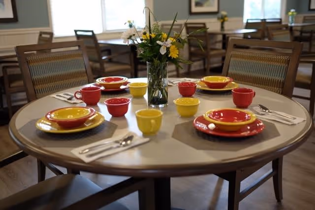 Round dining table set for four with red and yellow bowls, plates, and cups, silverware on napkins, and a vase of flowers in the center. Wooden chairs surround the table and more tables and chairs are visible in the background near windows.