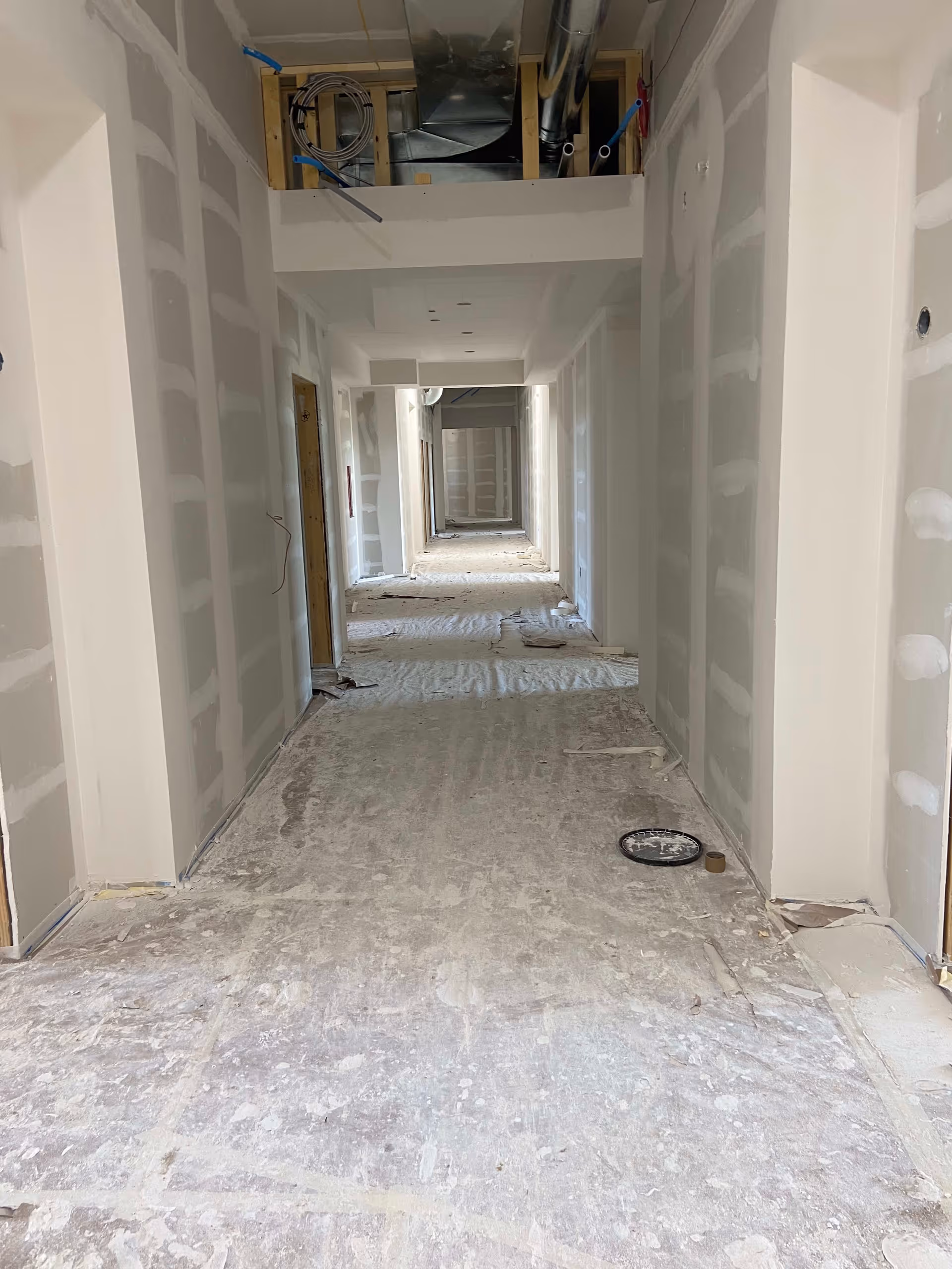 A long indoor hallway under construction with unfinished drywall on the walls and exposed ductwork and wiring on the ceiling. The floor is covered with dust and construction debris.