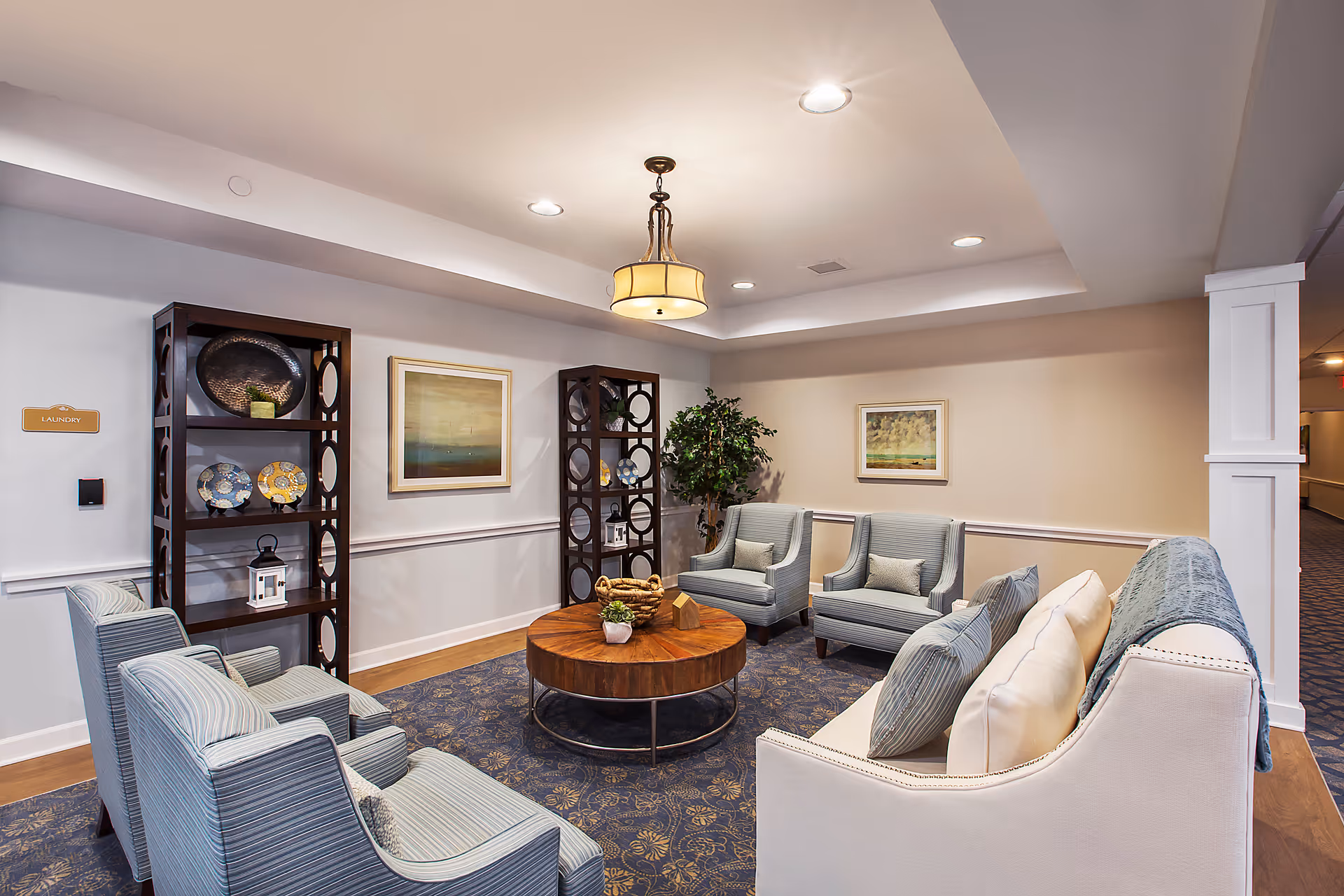 A cozy seating area in a senior living facility with a round wooden coffee table surrounded by six upholstered chairs and a sofa. Two dark wooden shelves with decorative plates and items flank a wall with two framed paintings. A potted plant is placed in the corner, and a ceiling light fixture illuminates the space.