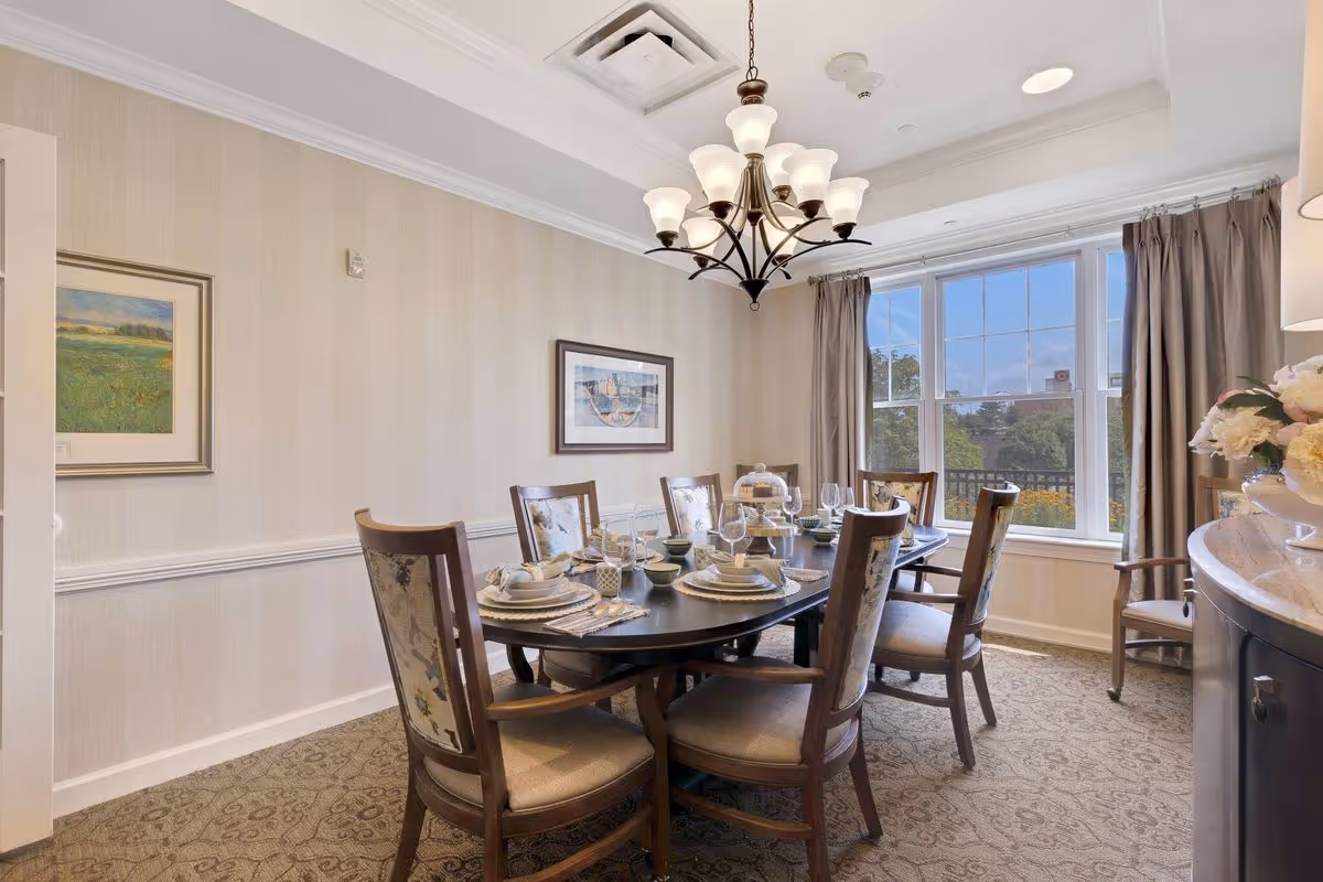 A well-lit dining room with a dark wooden oval table set for six people with plates, bowls, glasses, and napkins. The room has beige walls with two framed paintings, a large window with beige curtains letting in natural light, a chandelier hanging from the ceiling, and a sideboard with a floral arrangement.