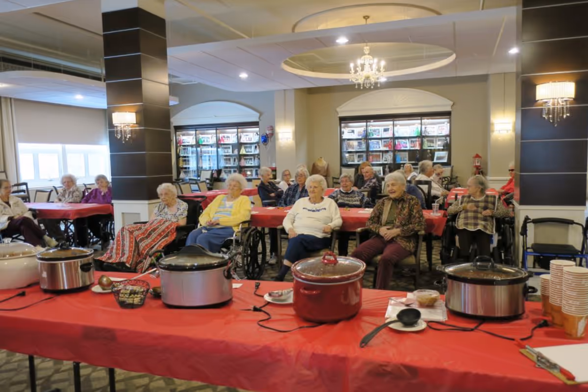 A group of elderly residents seated around tables covered with red tablecloths in a well-lit common room. Several slow cookers and cups are placed on a table in the foreground. The room has chandeliers, wall sconces, and large windows with shelves of books or magazines in the background.