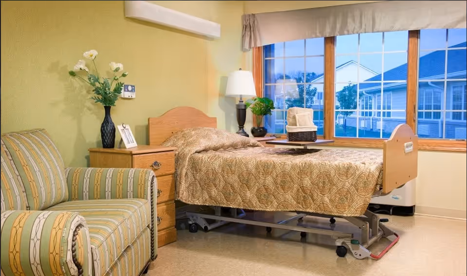 A single hospital-style bed with a patterned beige bedspread in a room with a large window showing an outdoor view. Next to the bed is a wooden nightstand with a black vase holding white flowers and a framed photo. A striped upholstered armchair is positioned to the left of the nightstand. A table lamp and a small plant are on a surface near the window.