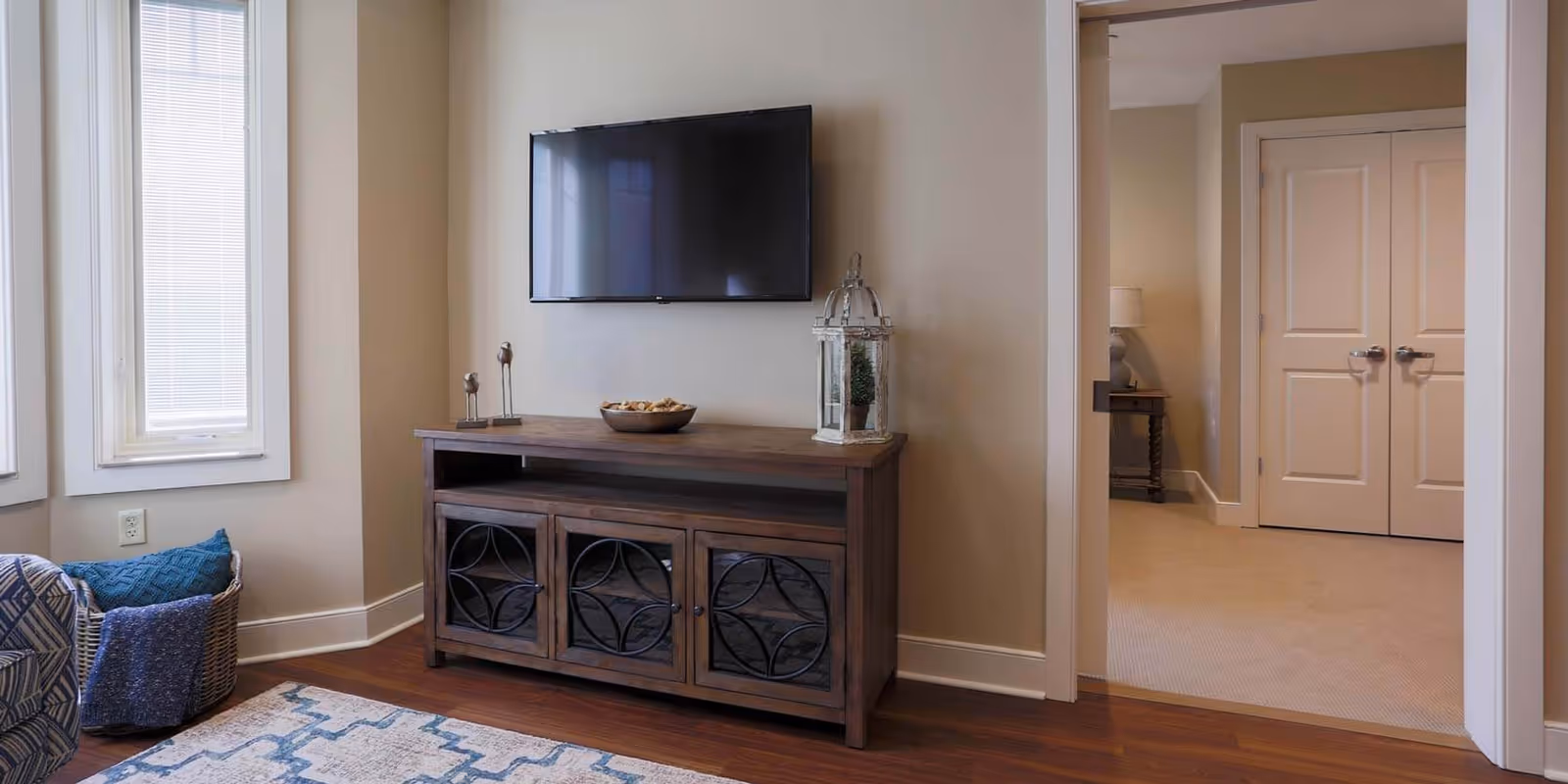 A cozy living room with a wall-mounted TV above a wooden console, decorative accents, windows, and a doorway to another room.