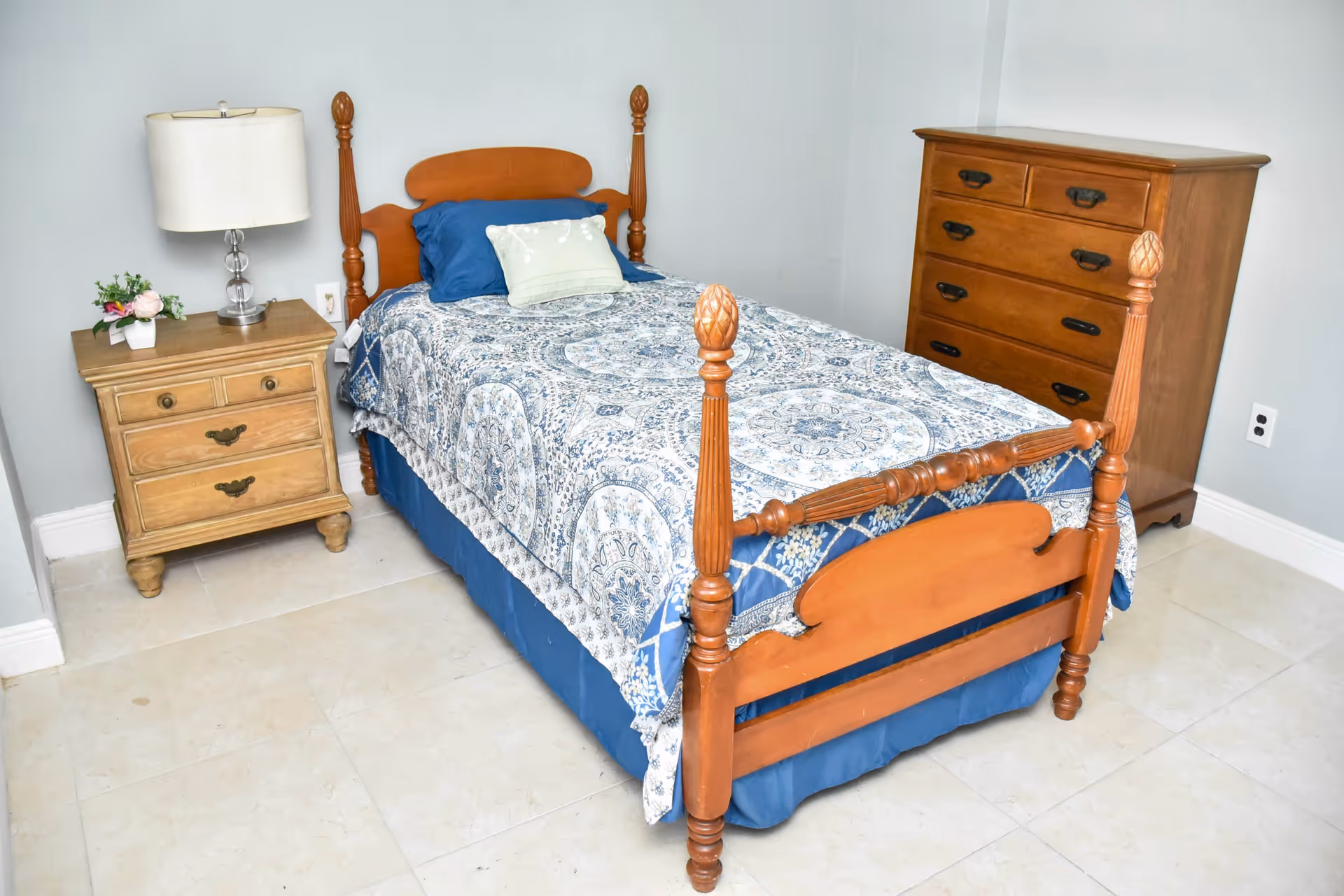 A small bedroom with a wooden single bed featuring a blue and white patterned bedspread and two pillows. Next to the bed is a wooden nightstand with a white lamp and a small flower arrangement. Across from the bed is a wooden chest of drawers. The room has light-colored tiled flooring and light gray walls.