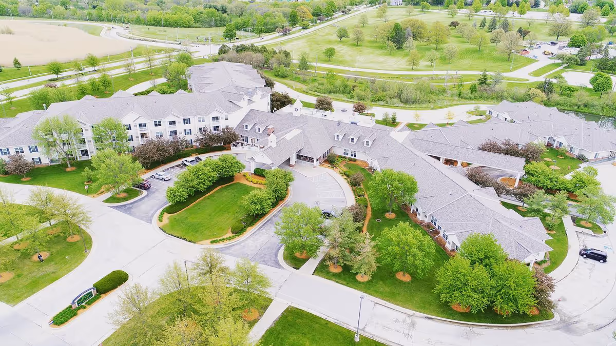 Aerial view of Melrose Meadows Retirement Community showing multiple connected buildings with gray roofs surrounded by green lawns, trees, and paved roads. The facility is situated near open fields and a park area with walking paths.