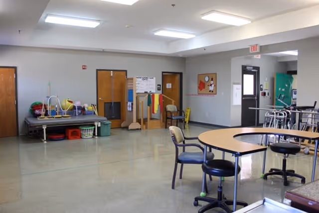 A spacious room with a polished floor, featuring a semi-circular table with chairs and stools around it. On the left side, there is a raised platform with various exercise and therapy equipment, including balls and hoops. The room has several wooden doors, a bulletin board, and an exit door with a window. The ceiling has recessed lighting panels.