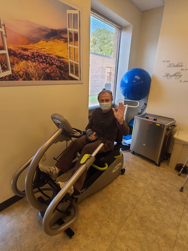 An elderly person wearing a face mask is sitting on a recumbent exercise bike in a room with a large window. The person is waving at the camera. The room has a large wall decal of an open window showing a scenic view of mountains and flowers, a blue exercise ball, and a small metal cabinet. There is also an inspirational quote on the wall.