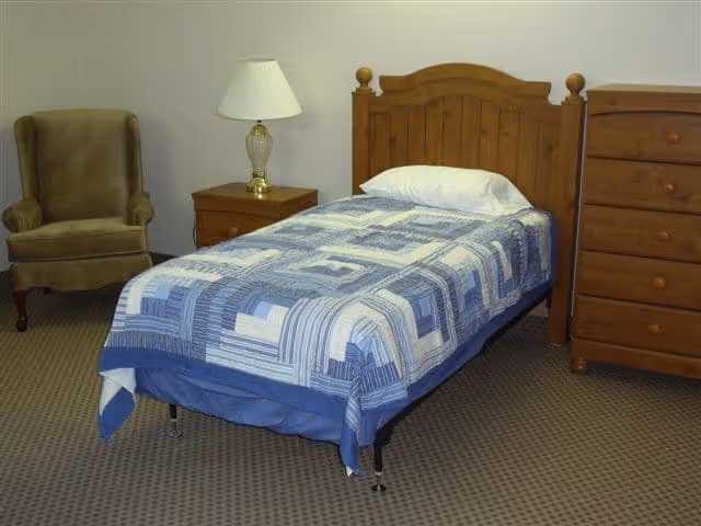 A simple bedroom with a single wooden bed featuring a blue and white patterned quilt and a white pillow. Next to the bed is a wooden nightstand with a glass lamp on it. To the left of the nightstand is a green upholstered armchair. On the right side of the bed is a wooden dresser with multiple drawers. The floor is carpeted in a neutral tone.