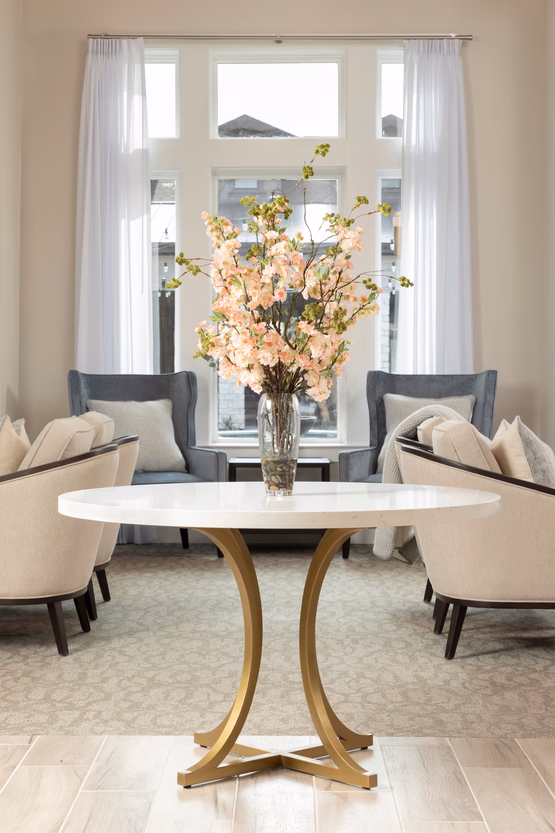Round table with a vase of pink blossoms in front of a large window flanked by cushioned armchairs.