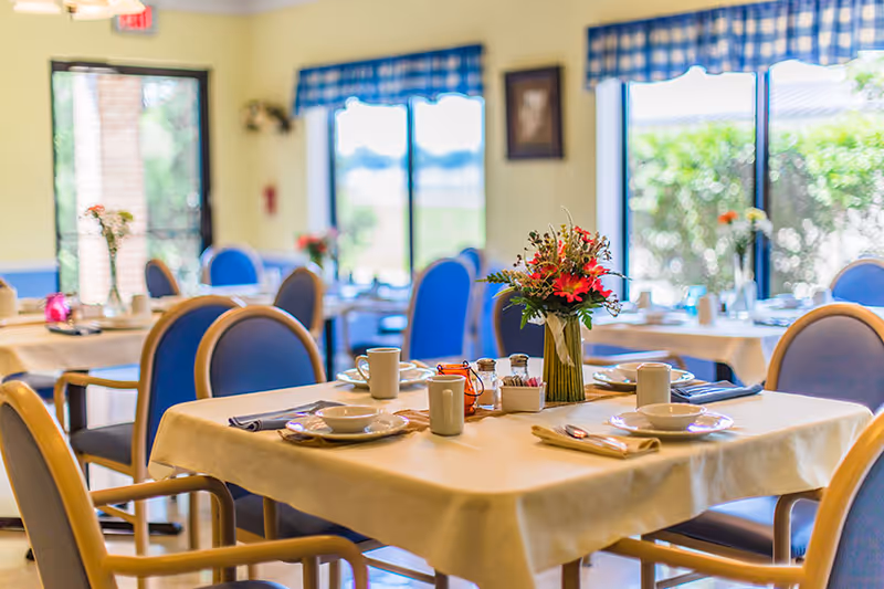 A bright dining room with tables covered in beige tablecloths, set with plates, bowls, cups, and utensils. Each table has a vase with colorful flowers. Blue cushioned chairs surround the tables. Large windows with blue checkered valances let in natural light and show greenery outside.
