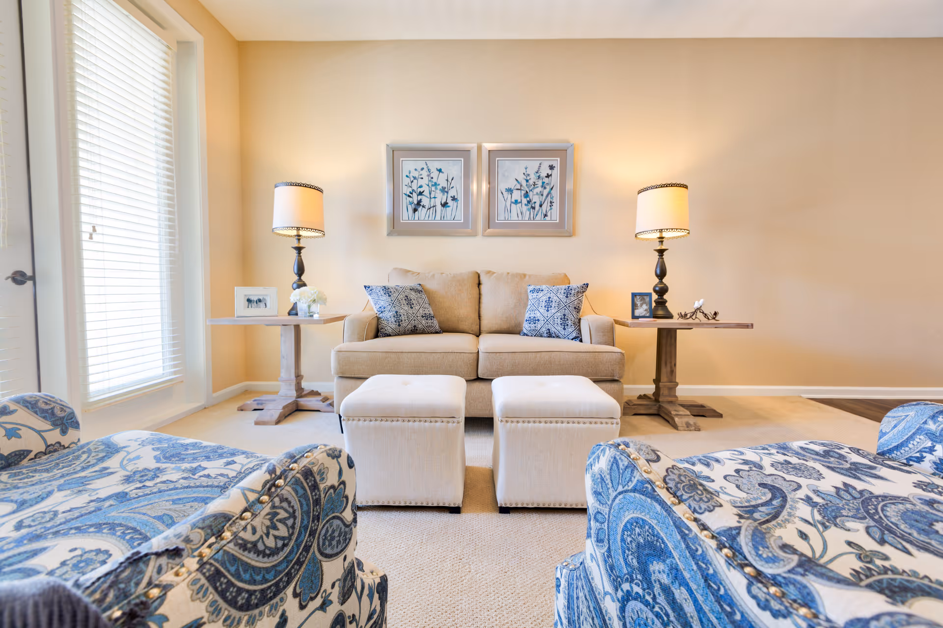 A bright living room with a beige sofa adorned with two blue patterned pillows, flanked by two wooden side tables each holding a lamp and decorative items. In front of the sofa are two white ottomans, and two blue patterned armchairs are partially visible in the foreground. The walls are light beige with two framed floral artworks hanging above the sofa. Large windows with blinds allow natural light to fill the room.