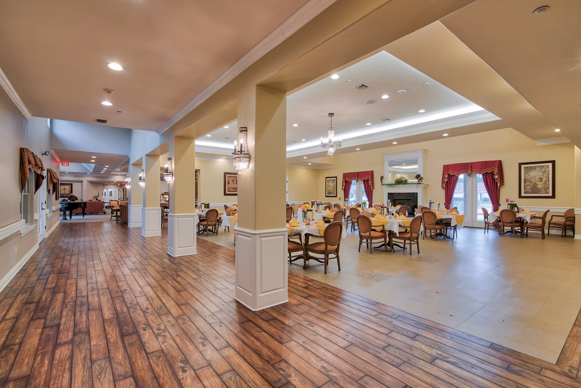 Interior view of the dining room at Pelican Bay Assisted Living Community, featuring tables set with yellow tablecloths and chairs, a fireplace with decorative items, and elegant lighting fixtures.