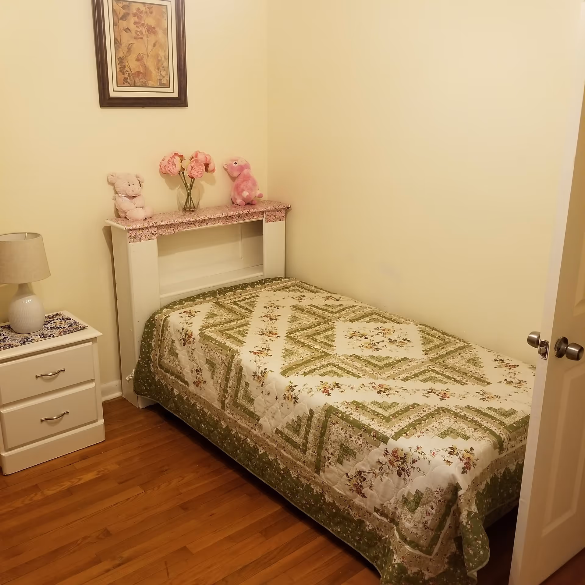 A small bedroom with a single bed covered in a green and white floral quilt. There is a white nightstand with two drawers and a lamp on top. Above the bed is a framed floral picture on the wall. On the headboard shelf, there are two stuffed animals and a vase with pink flowers. The floor is wooden, and the walls are painted light yellow.
