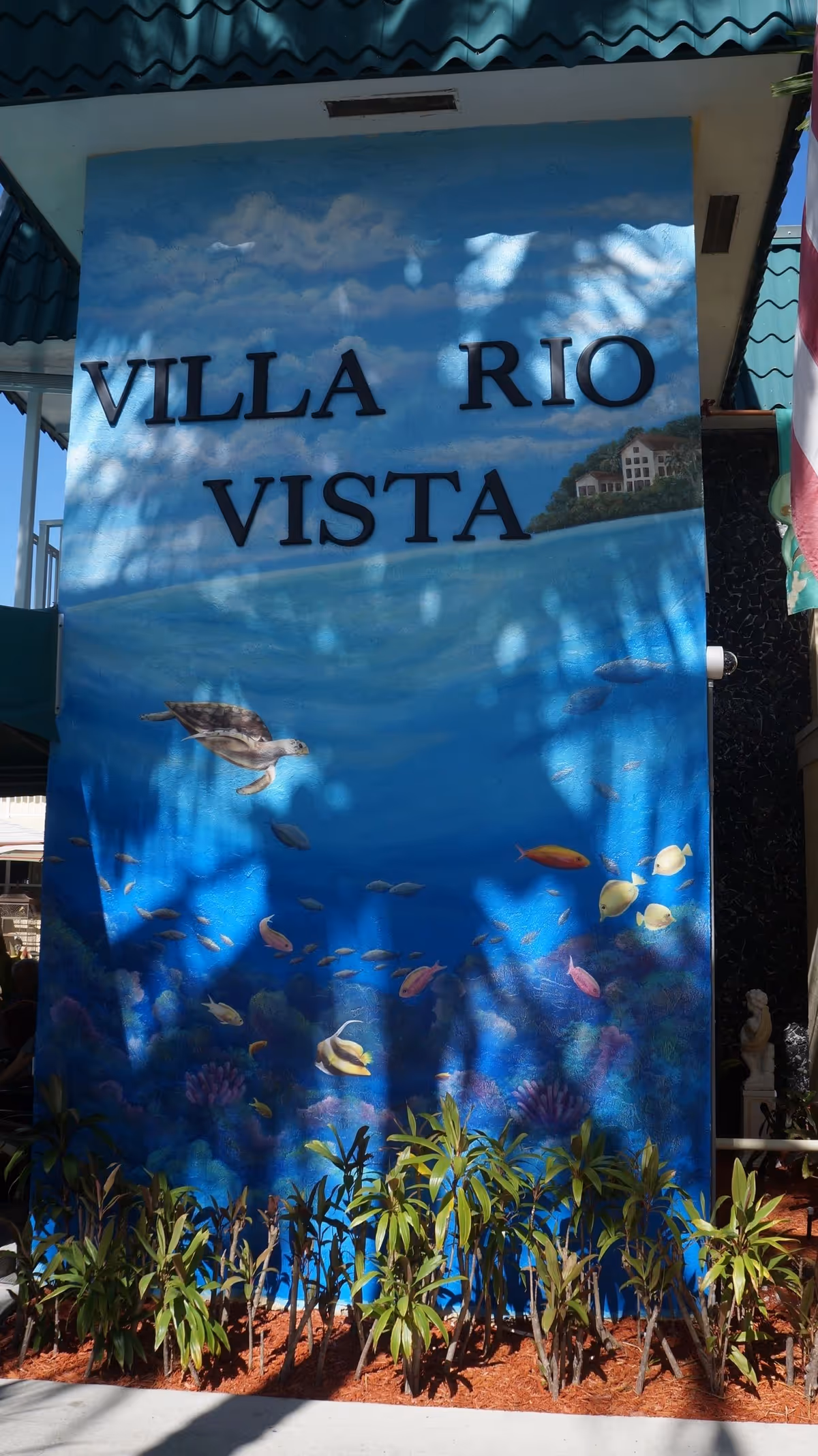 A mural painted on a wall with the text 'VILLA RIO VISTA'. The mural depicts an underwater scene with various fish and a sea turtle swimming, along with a small house on a hill in the background. There are plants growing at the base of the wall and shadows of tree branches cast on the mural.