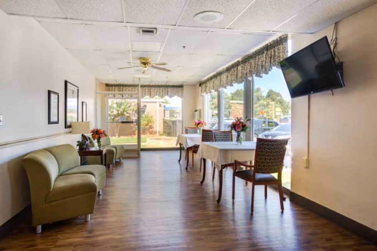 A bright interior room with wooden flooring featuring two green armchairs on the left side, a small table with a lamp and flowers between them, and two tables with white tablecloths and floral centerpieces near large windows on the right. A wall-mounted TV is visible on the right wall, and outside the windows, cars and greenery can be seen.
