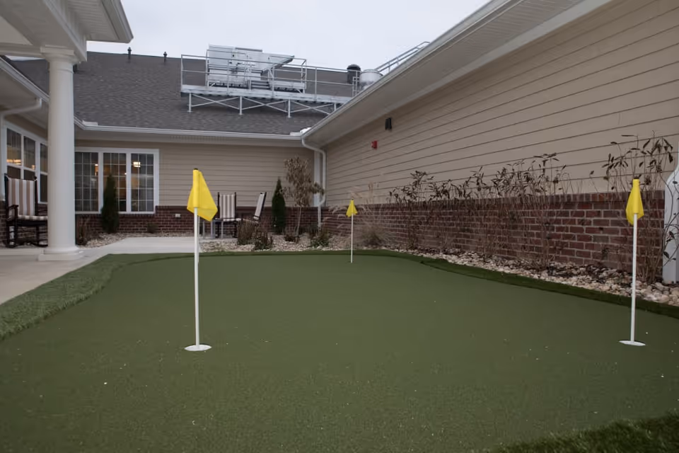 A small outdoor putting green with three yellow flags is situated in a courtyard area surrounded by beige siding walls and a brick lower wall. There are patio chairs and small tables near the windows of the building, and some landscaping with small bushes and rocks along the edges.