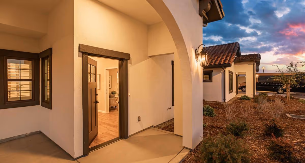View of the exterior entrance to a senior living facility with an open door leading inside. The building features white walls, dark trim around windows and doors, a tiled roof, outdoor lighting fixtures, and landscaped plants. The sky is partly cloudy with a colorful sunset.