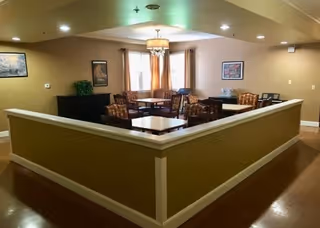 Interior view of a common area with multiple square tables and cushioned chairs arranged around them. The space has beige walls, wooden flooring, framed artwork on the walls, and a window with curtains letting in natural light. A chandelier-style light fixture hangs from the ceiling.