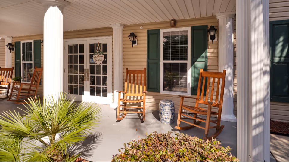 Covered front porch with wooden rocking chairs, white columns, potted plants and double glass entry doors.