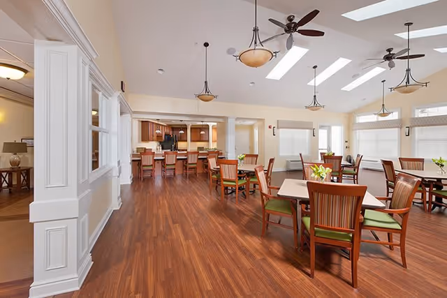 A spacious dining room with wooden floors, several tables and chairs with green cushions, ceiling fans, pendant lights, and large windows with blinds. In the background, there is a kitchen area with a counter and bar stools.