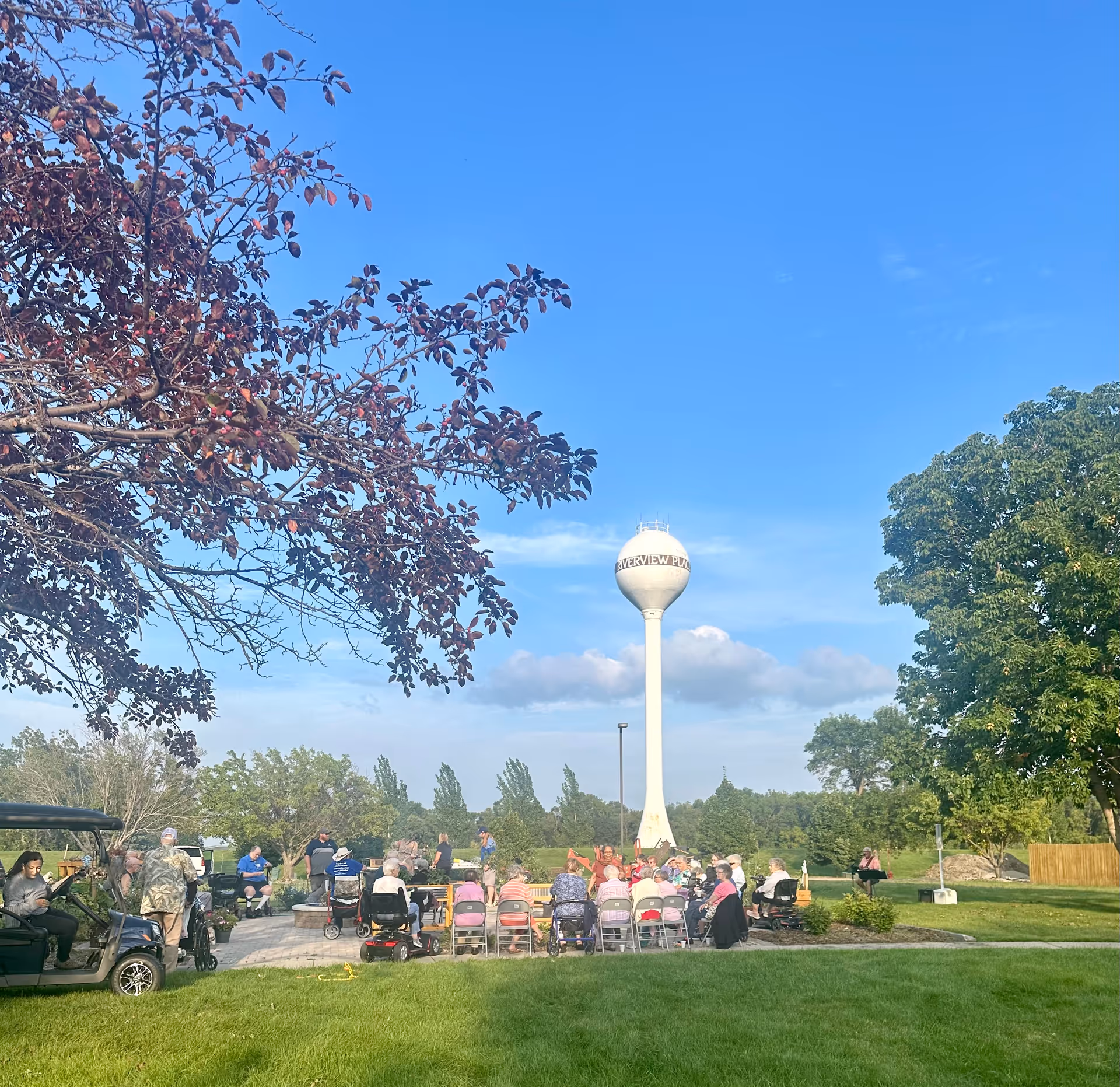 A group of elderly people seated outdoors in a park-like setting with green grass and trees, facing a water tower labeled 'Riverview Place' under a clear blue sky.