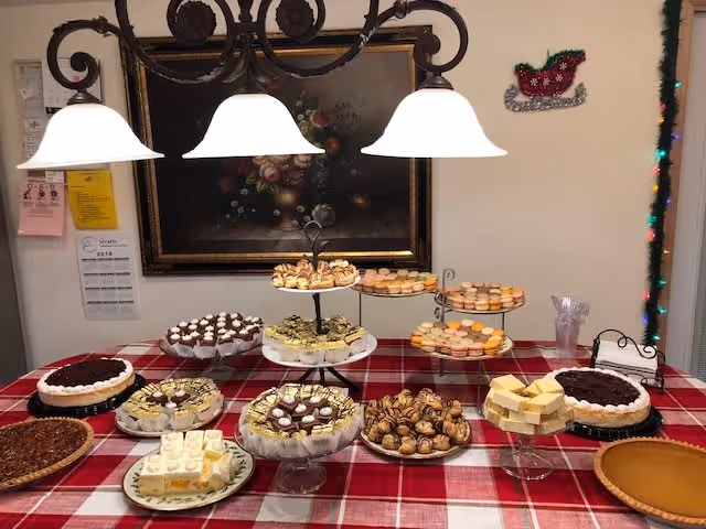 A dessert table covered with a red and white checkered tablecloth displaying various cakes, pastries, and cookies under three hanging white lamps. A framed floral painting hangs on the wall behind the table, with a small decorative sleigh on the right wall and a string of colorful lights along the door frame.
