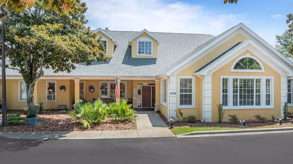 Front entrance of a yellow senior living building with a covered porch, landscaping, and an American flag.