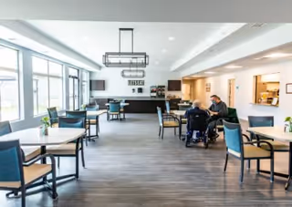 A bright dining room in a senior living facility with multiple tables and chairs arranged neatly. Large windows on the left side let in natural light. Two people are seated at a table on the right side, one in a wheelchair. The back wall has a counter area with coffee machines and a sign that reads 'Serene Gardens of Clarkston'.