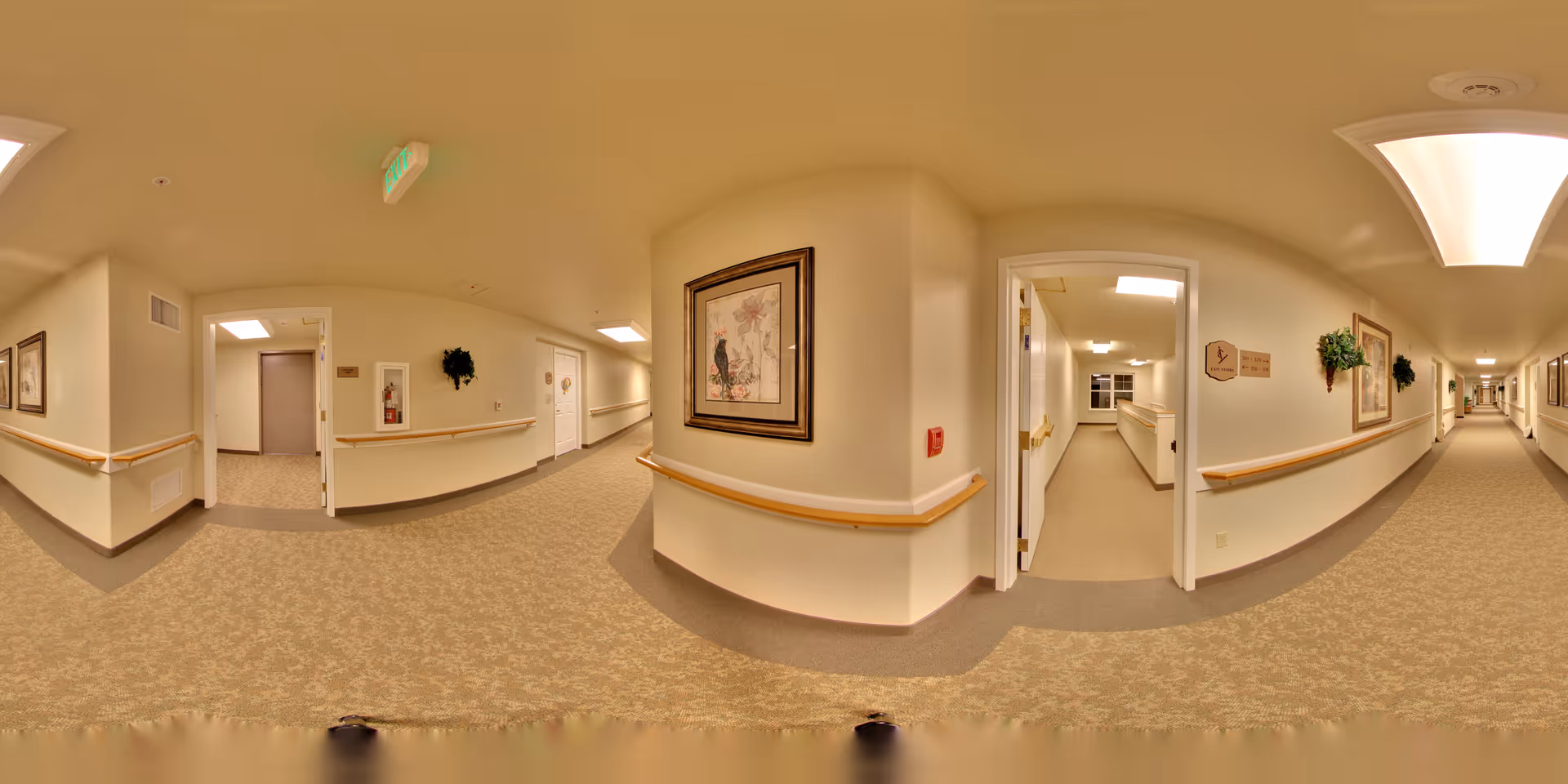 A wide hallway inside a senior living facility with beige walls and carpeted floors. The hallway has handrails on both sides, framed artwork, and some green wall decorations. There are several doors along the hallway, one of which is open revealing another corridor. The lighting is bright with ceiling lights, and an exit sign is visible above one door.