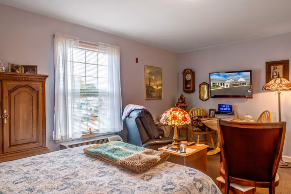 A cozy bedroom in a senior living facility with a bed covered in a patterned quilt and folded blankets. Next to the bed is a black recliner chair and a wooden side table with a decorative lamp. The room has a window with white curtains, a wooden cabinet, a wall clock, and a small table with chairs. A flat-screen TV is mounted on the wall displaying an image of the facility exterior.