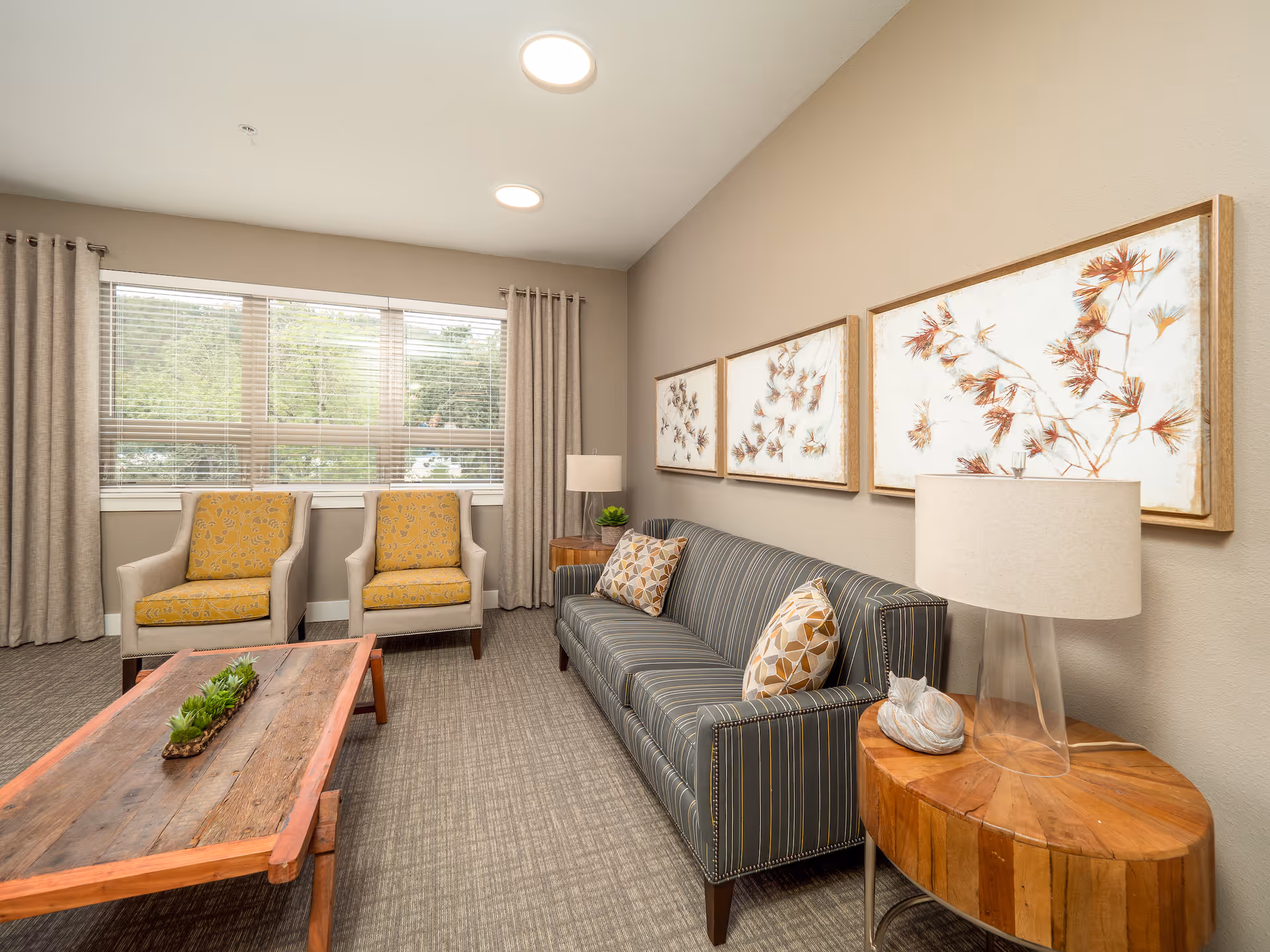 A cozy living room with a striped sofa adorned with patterned cushions, two beige armchairs with yellow patterned cushions, a wooden coffee table with a small succulent centerpiece, and a round wooden side table with a clear lamp and decorative item. Three framed botanical prints hang on the beige wall, and large windows with beige curtains allow natural light to fill the room.