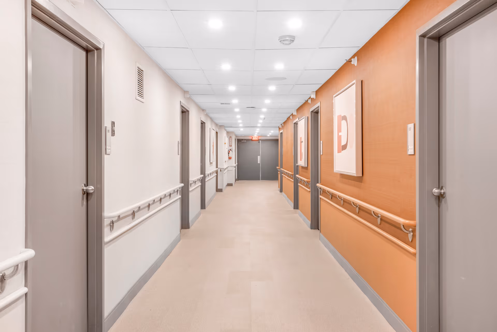 A clean, well-lit hallway in a healthcare facility with beige and orange walls, multiple closed gray doors on both sides, handrails along the walls, ceiling lights, and framed artwork on the right wall.