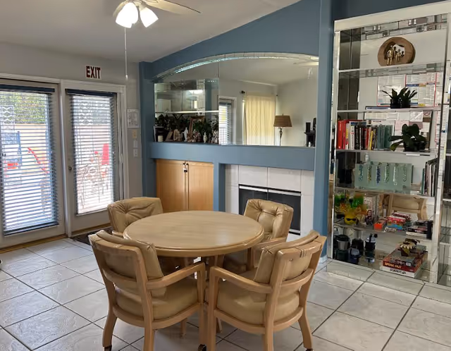 A cozy interior room with a round wooden table surrounded by four cushioned chairs. The room features a fireplace with a blue mantel, a large mirror above it, and a built-in glass shelving unit filled with books and decorative items. There are double glass doors with blinds leading outside, and a ceiling fan with lights overhead.