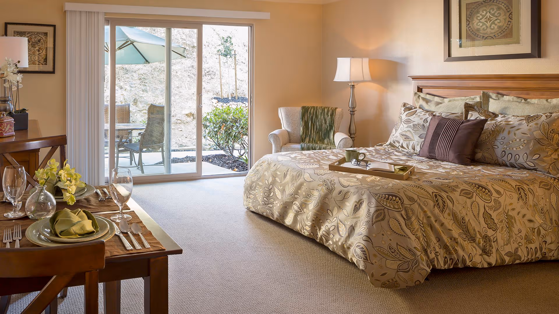 A cozy bedroom with a large bed covered in patterned bedding and multiple pillows. A wooden tray with a cup and book rests on the bed. To the right of the bed is a cushioned armchair with a green throw and a floor lamp. On the left side, there is a dining table set for two with plates, glasses, and napkins. Sliding glass doors lead to an outdoor patio with chairs and an umbrella. The room is warmly lit and decorated with framed artwork on the walls.