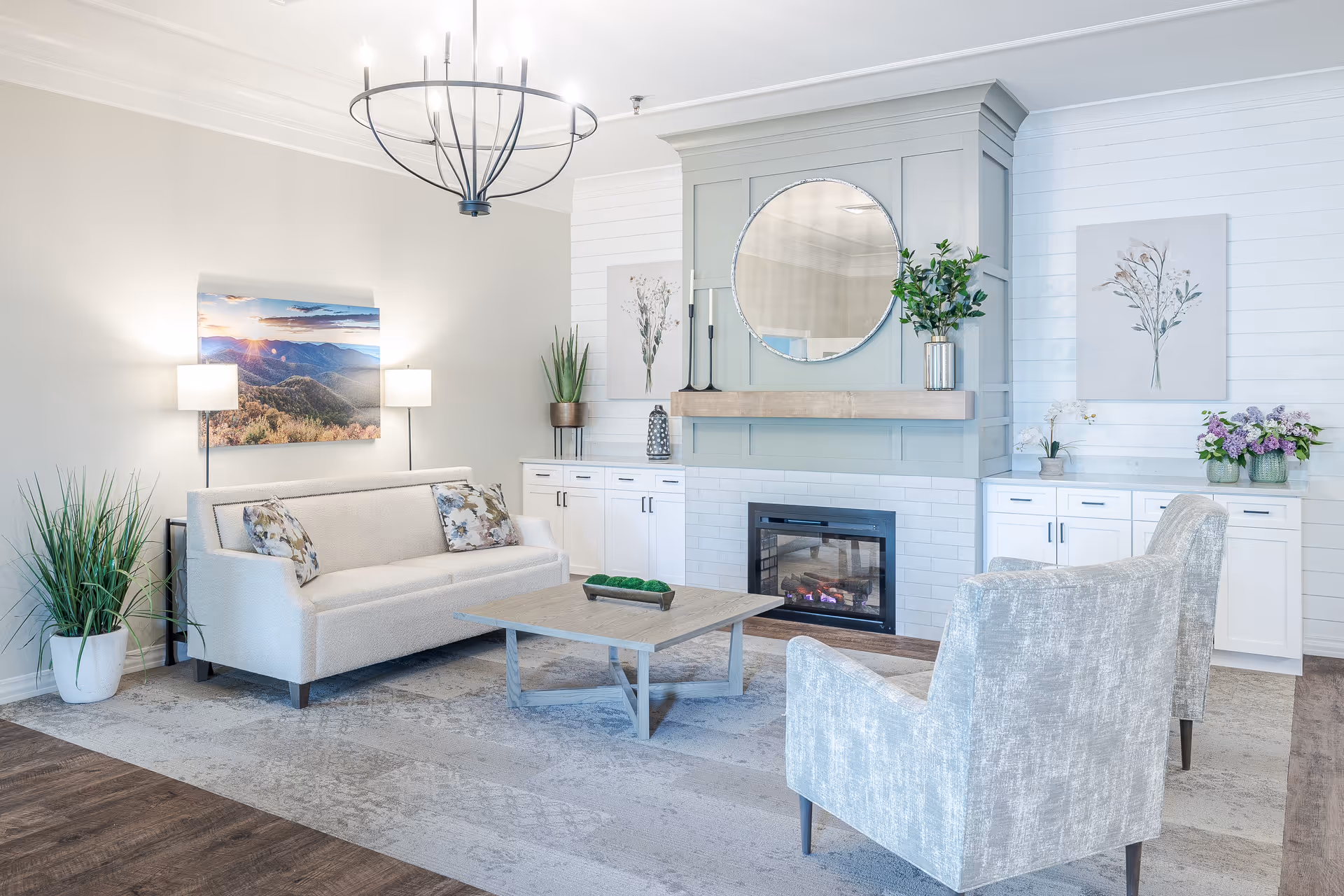 A bright and modern living room with a beige sofa, two light gray armchairs, a wooden coffee table, and a fireplace with a round mirror above it. The room features white cabinetry, decorative plants, floral artwork, and a chandelier hanging from the ceiling.