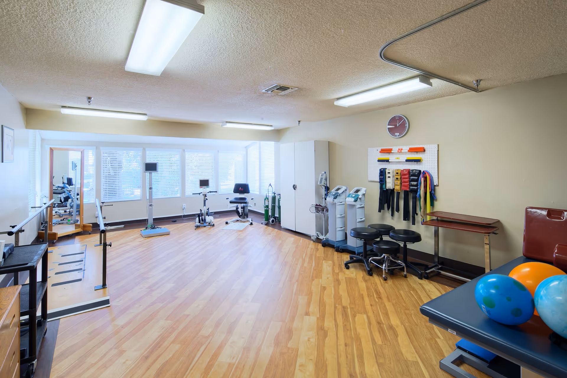 A spacious physical therapy or rehabilitation room with wooden flooring, exercise equipment including stationary bikes, parallel bars, therapy balls on a padded table, resistance bands hanging on a pegboard, stools, and medical devices. The room is well-lit with fluorescent ceiling lights and has large windows with blinds allowing natural light.
