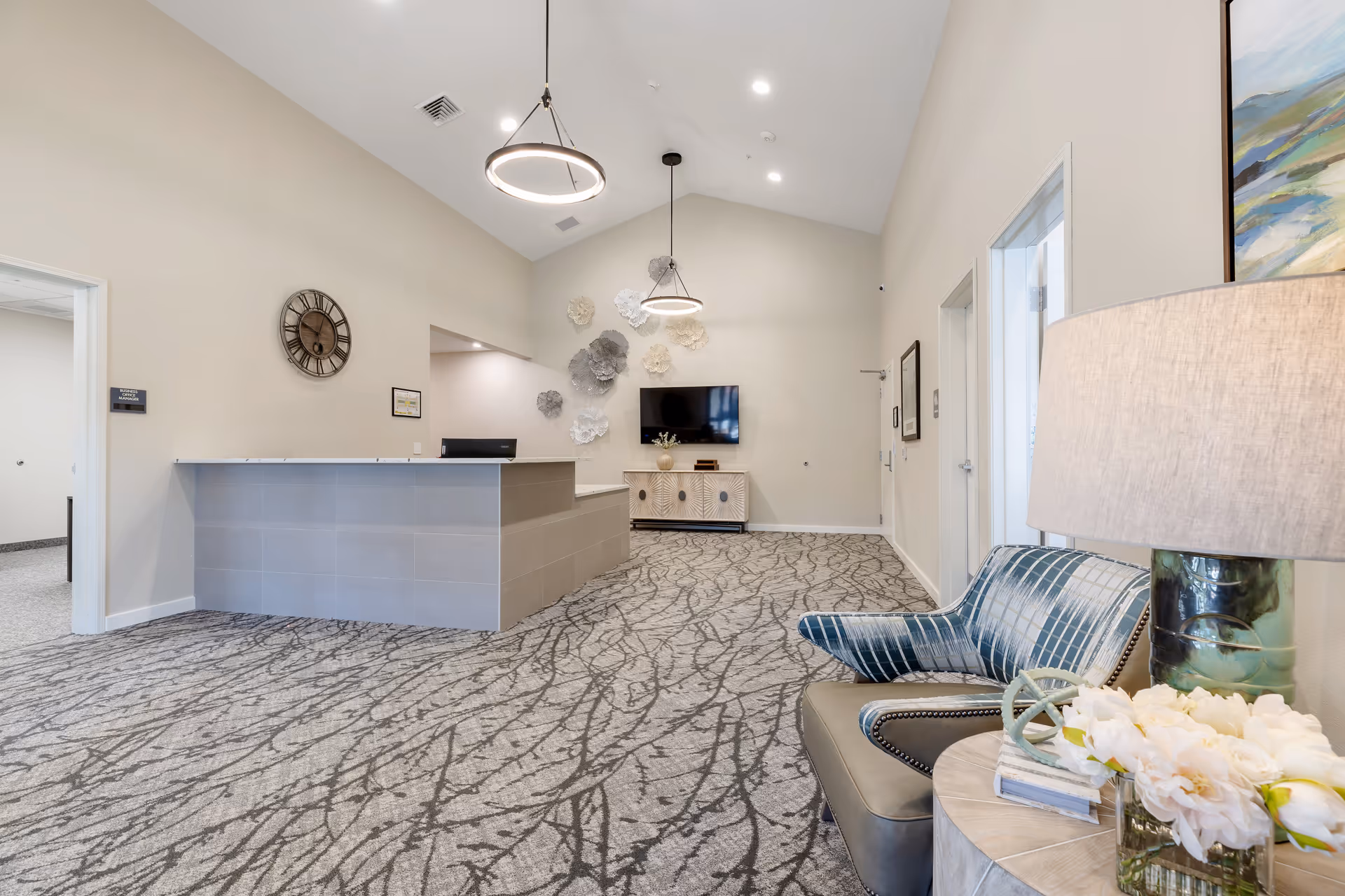 Reception area of White Oak Village Memory Care with a modern front desk, patterned carpet, a wall clock, decorative wall art, a flat-screen TV mounted on the far wall, and a seating area with a blue plaid armchair and a side table with a lamp and flowers.