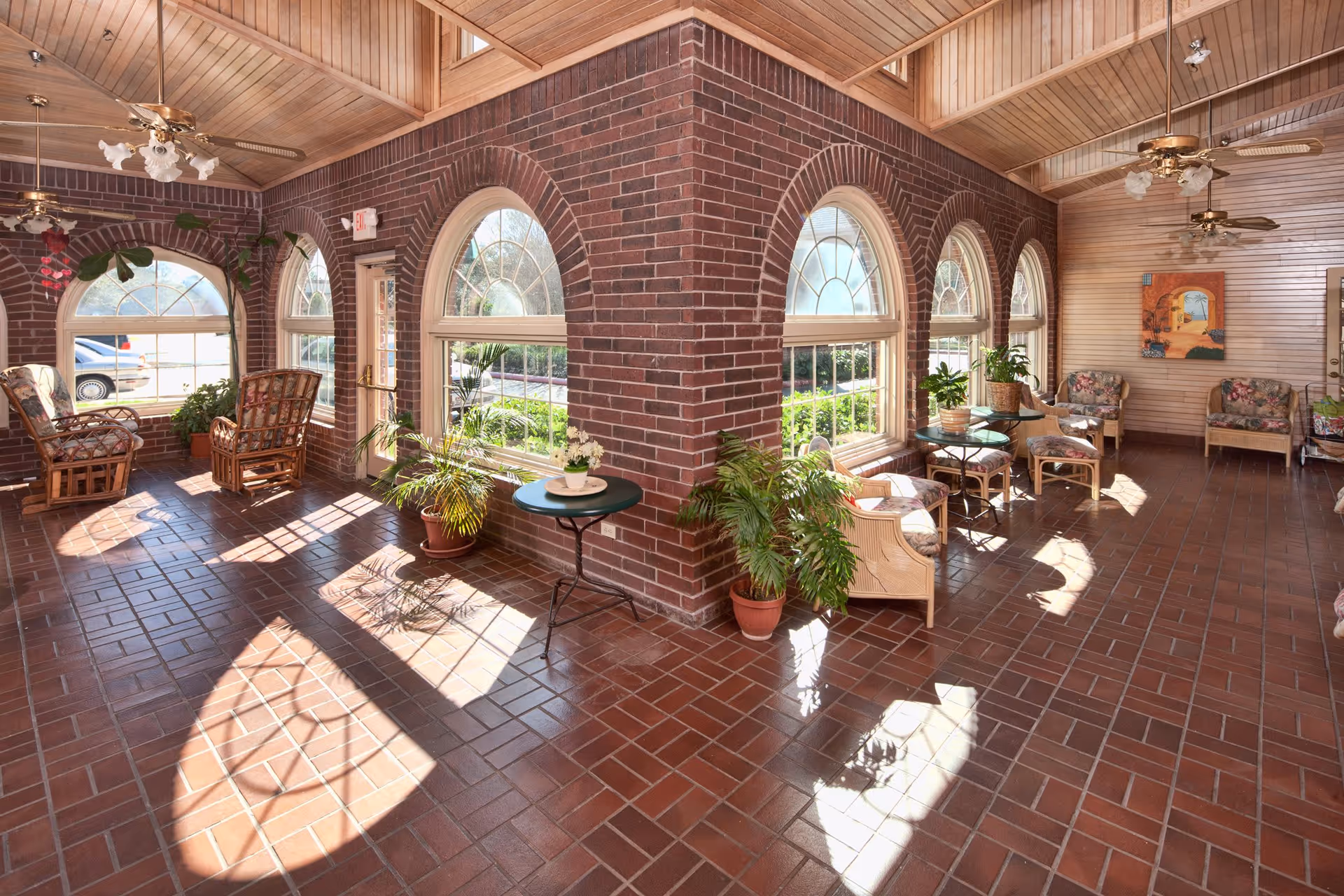 A sunlit interior common area with brick walls and large arched windows. The room features several wicker chairs with floral cushions, small tables, potted plants, ceiling fans, and a wooden ceiling. Sunlight casts shadows on the tiled floor.