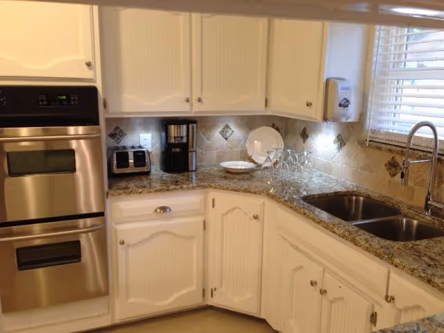 A clean and modern kitchen corner with white cabinets, granite countertops, a double stainless steel sink with a high-arc faucet, a built-in stainless steel oven, a coffee maker, a toaster, and a window with white blinds letting in natural light.
