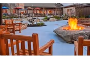 Outdoor seating area with wooden chairs arranged around a large stone fire pit with a fire burning. In the background, there are more seating areas with umbrellas and a building structure surrounding the courtyard.