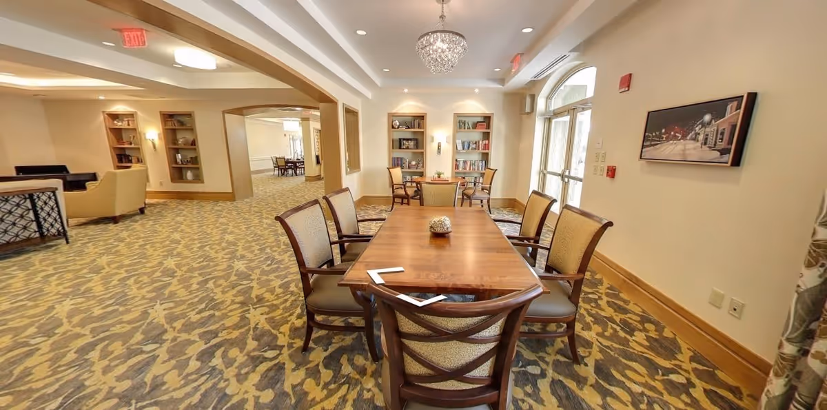 A spacious and well-lit common area with a wooden table surrounded by six cushioned chairs. The room features patterned carpet flooring, built-in bookshelves with books and decorative items, a chandelier hanging from the ceiling, and large windows letting in natural light. There is a painting on the wall and additional seating areas visible in the background.